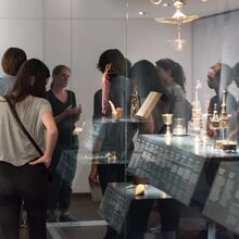 Foto eines Ausstellungsbereichs. Um eine große Vitrine mit metallischen Gegenständen und einem Buch ist eine Gruppe versammelt, die eine Führung erhält.
