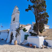 Foto eines weißen Gebäudes im griechischen Stil vor blauem Himmel. Über einer Tür ist ein Turm mit einer Glocke. Vor dem Gebäude ist ein Natursteinboden. Auf dem Gebäude weh eine griechische Flagge.