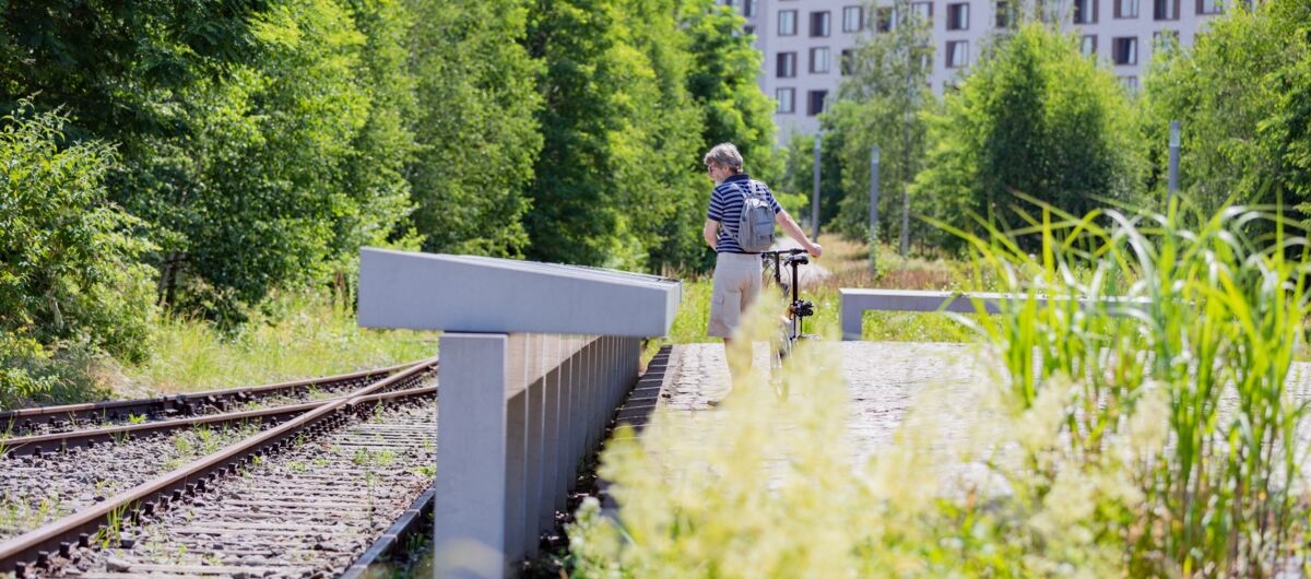 Blick aus Kniehöhe. Links sind zwei Gleise zu sehen. Mittig eine lange Reihe von Informationstafeln aus Beton, die von einem Menschen mit Fahrrade betrachtet werden. Im Hintergrund sind grüne Bäume sowie eine Reihenhauszeile zu sehen.