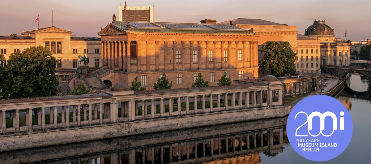 Foto der Museumsinsel Berlin. Im Vordergrund ist die Spree. Dahinter sind die Alte Nationalgalerie, Teile des Pergamonmuseums und des Neuen Museums sowie des Kolonnadenhofs zu sehen -  alles alte, prächtige und aus Stein errichtete Bauwerke.