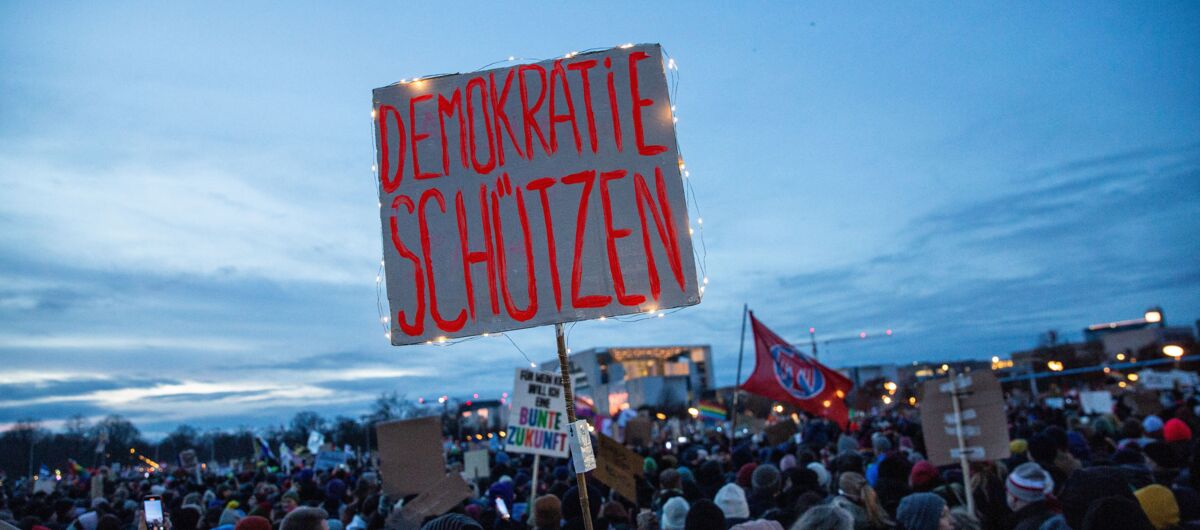 Demonstrierende Menschenmenge. Viele halten Schilder hoch. Eines sticht besonders heraus, da es mit einer Lichterkette umwickelt ist und in der aufkommenden Dämmerung, die im Hintergrund aufzieht, leuchtet. Auf dem Schild steht "Demokratie schützen".