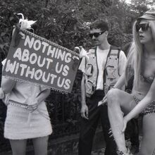 Schwarz-weiß Foto mit drei Personen im Freien. Die linke Person scheint eine Frau zu sein. Sie hält ein Schild mit der Aufschrift "Nothig about us without us" vor ihr Gesicht. Mittig steht ein Mann. Rechts eine Frau im Bikini, die sich ein Bein eincremt.