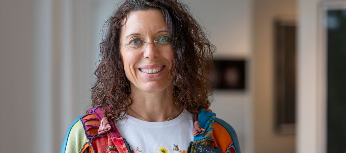 Foto in einem Ausstellungsraum. Eine freundlich lachende Frau ist bis zum Oberkörper erfasst. Sie trägt schulterlanges lockiges dunkles Haar, eine unscheinbare Brille, ein weißes T-Shirt mit Blumenmotiv und darüber eine bunte Patchwork-artig Kapuzenjacke.