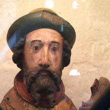 Foto des Oberkörpers einer Männerfigur mit grüner Kopfbedeckung und braunem Bart. Es wurde durch eine Glasscheibe, die durch Eisenstangen geziert wird, hindurchfotografiert.
