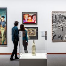 Foto eines Ausstellungsbereichs. Im Vordergrund mittig steht in einer Vitrine eine metallisch wirkende Coca-Cola-Flasche. Dahinter sind vier Gemälde unterschiedlicher Form und Stil, die von zwei Menschen betrachtet werden.