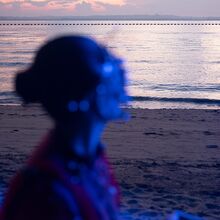 Foto, das im Vordergrund einen blau angeleuchteten Oberkörper einer Person zeigt. Er ist verschwommen. Der Hintergrund ist scharf. Zu sehen ist in dämmeriger Stimmung ein Strand. Das Meer ist ruhig. Wolken sind am Himmel.