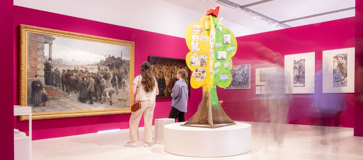 Foto eines Ausstellungsbereichs. An einer roten Wand hängen große und kleine Gemälde, die vornehmlich Menschengruppen abbilden. Zwei Frauen sehen sich die Bilder an. Hinter ihnen steht auf einem runden Podest ein gebastelter Baum.