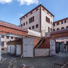 Außenansicht der Gedenkstätte Lindenstraße. Das verschachtelte Backsteingebäude ist teils weiß verputzt. Der Boden ist aus Kopfsteinpflaster. Im Vordergrund sind rechts und links zwei Durchgäng mit Flügeltüren zu sehen, die zu dem Hauptgebäude führen.