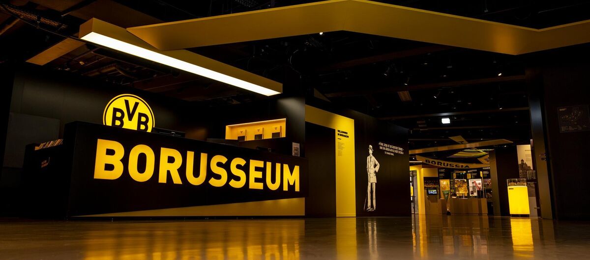 Vom Boden aufgenommenes Foto. Links eine Theke, auf der "Borusseum" steht. Dahinter das BVB-Logo. Rechts gehen die Ausstellungsräume ab. Die Vereinsfarben werden durch schwarze Wände und gelbe Buchstaben, Logos und Ausstellungsinhalte wiedergespiegelt.
