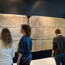 Foto von drei Menschen, die vor einer schwarzen Wand mit hellen großen Steinplatten stehen. In diesen sind verschiedene Szenen großflächig hineingemeißelt. Die Steinplatten haben ein Querformat und sind in drei Reihen übereinander angeordnet.