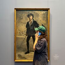 Foto eines Menschen mit langen lockigen Haaren, weitem Hemd und Kappe vor einem Gemälde. Die Person hält ein Handy an das Ohr während sie sich das Gemälde anschaut.