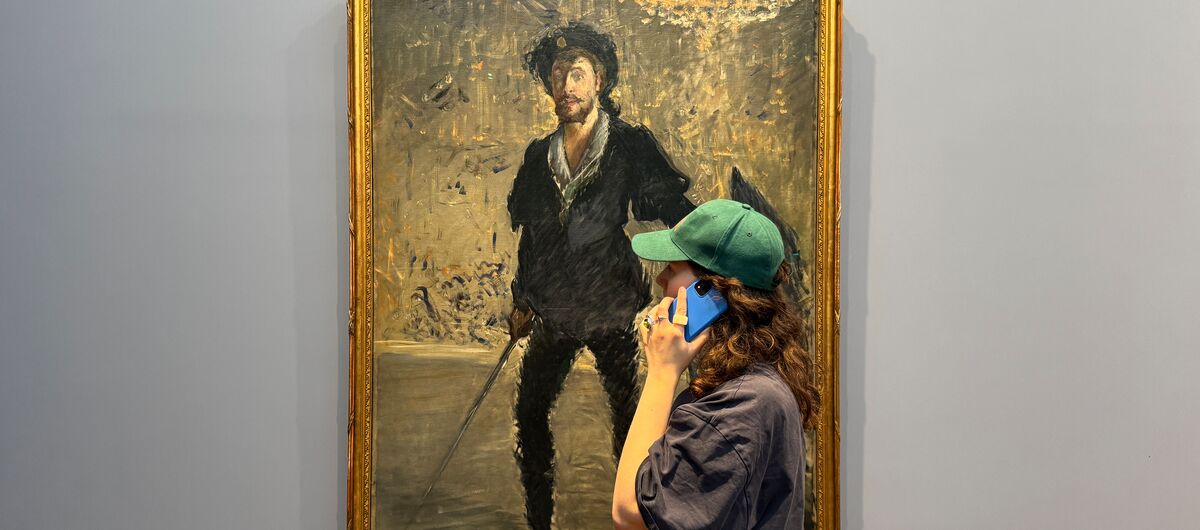 Foto eines Menschen mit langen lockigen Haaren, weitem Hemd und Kappe vor einem Gemälde. Die Person hält ein Handy an das Ohr während sie sich das Gemälde anschaut.