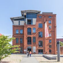 Eingang der "Neuen Fabrik", dem Gebäude des Museums der Arbeit. Es besteht aus rotem Backstein und großen Bogenfenstern mit Sprossen. Neben dem Haus steht ein hoher Schornstein aus Backstein.