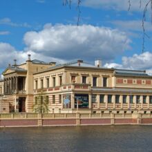 Seitlicher Blick auf das Staatliche Museum Schwerin. Im Vordergrund ist ein See. Das Gebäude ist sandsteinfarbig und hat viele Fenster. Auf der linken Seite des Fotos ist der Eingang zu sehen, vor dem sechs dunkele Säulen stehen. Zu ihm führt eine Treppe.