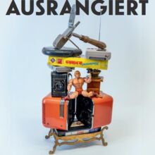 Gezeigt wird das Werbeplakat zur Sonderausstellung "Ausrangiert". Gestapelt sind alte Gegenstände. Von unten nach oben: Tisch, Videokassette, Aktionfigur, Fotoapparat, Kaffeemühle, Buch, CD-Player, Gameboy Klapphandy in einem Art Selfistick.