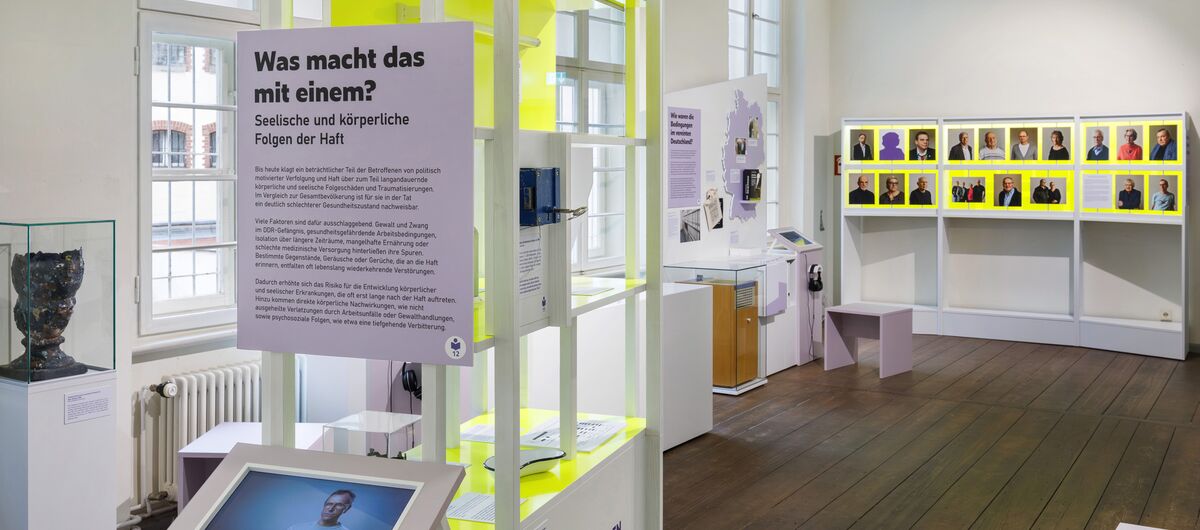Foto eines Ausstellungsbereichs. Auf dunklem Holzboden stehen moderne Vermittlungsstationen in Form von Bildschirmen mit Kopfhörern, Vitrinen, Texttafeln sowie 19 kleine drehbare Tafeln mit Foto auf der Vorder- und Text auf der Rückseite.