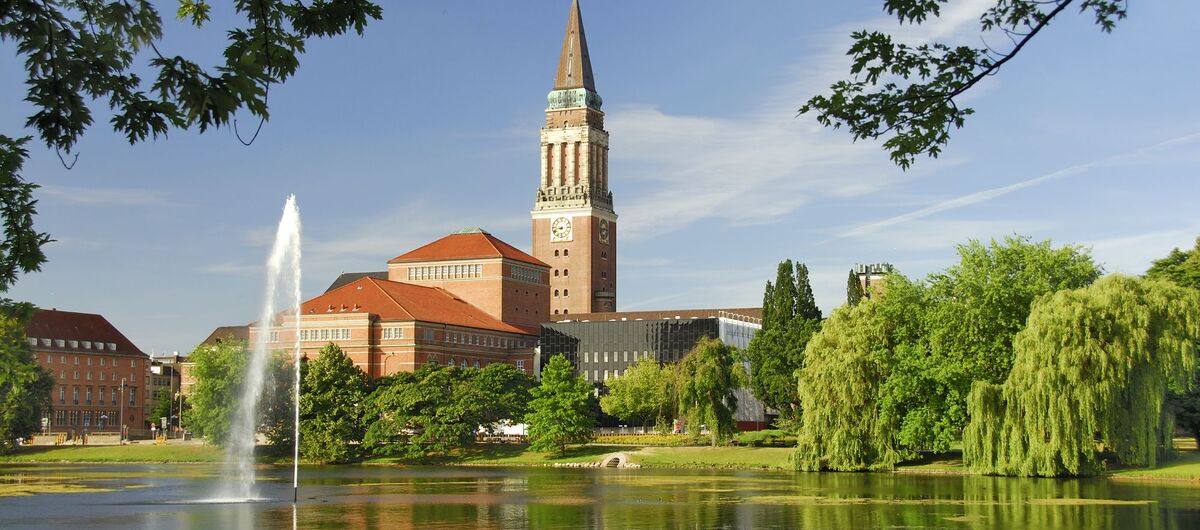 Foto des Kieler Rathauses. Im Vordergrund ist ein See mit Enten und Wasserfontäne. Dahinter schließt ein Grünsteifen mit Wiese und Bäumen an. Im Hintergrund sind die Gebäude zu sehen. Ein hoher Turm ragt mittig hervor.