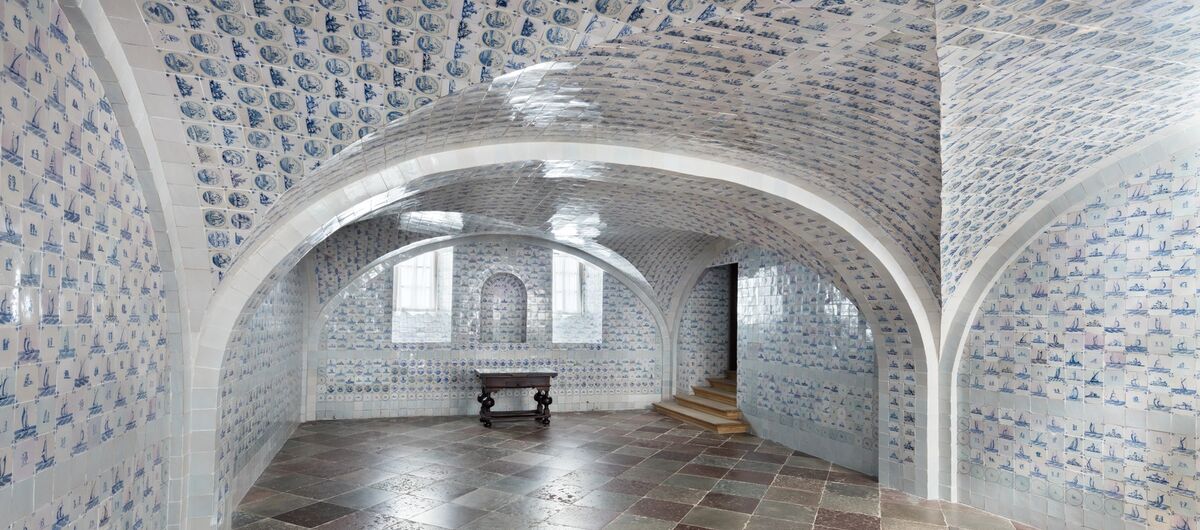 Gewölberaum, der von oben bis unten mit Fliesen ausgekleidet ist. Zwei Fenster sind in Nischen eingelassen. Durch die blau-weißen Fliesen ist der Raum hell, wirkt aber kühl. Ein kleiner dunkler Tisch steht an einer Wand. Stufen führen rechts aus dem Raum.