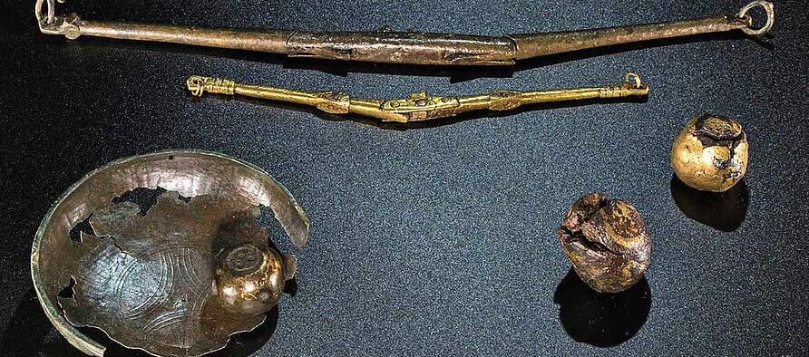 Foto metallischer Teile von Klappwaagen mit Schalen und Gewichten auf schwarzem Untergrund. Oben liegen eine große und eine kleine Klappwaage , links eine löchrige Schale mit einem Gewicht, daneben zwei weitere kugelförmige Gewichte.
