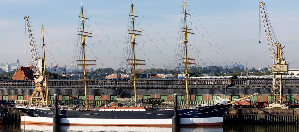 Der Frachtsegler Peking liegt im Hafen vor Anker. Es ist ein vier Masten tragendes Schiff mit insgesamt 32 Segeln, die eingeholt sind. Rechts und links des Schiffes steht je ein Hafenkran. Es ist ein freundlicher Tag.