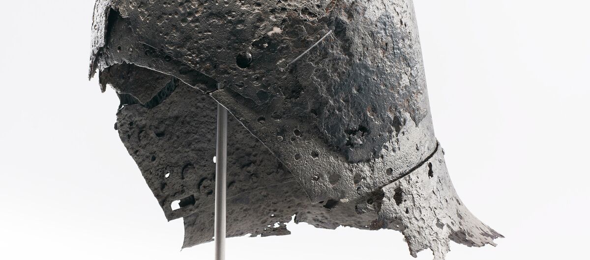 Foto einer historischen Beckenhaube. Sie ist auf einen Stab gesetzt, der in einen hellen Holzsockel mündet. Der eiserne Helm weist eine unebene Oberfläche auf. Teilweise Durchziehen ihn Löcher. Die Ränder sind ausgefranst.