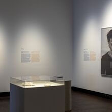 Foto eines Ausstellungsbereichs. An der rechten Wand hängt ein großes Portraitgemälde eines Mannes, an der linken Wand hängt ein kleines Gemälde - beide um Texte ergänzt. Im Raum steht eine Vitrine mit zwei Exponaten, die erleuchtet sind.