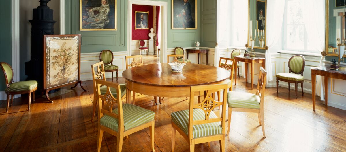 Foto eines barocken Zimmers mit Holzboden. Die Wände, an denen Gemälde hängen, sind weiß und grün gestrichen. In der Mitte steht ein runder Holztisch mit sieben Holzstühlen, deren Sitzflächen grün-weiß gestreift bezogen sind.