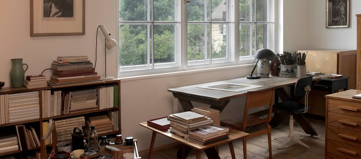 Foto eines Arbeitszimmers mit Schreibtisch und zwei Stühlen vor einem großen Sprossenfenster mit Blick auf einen bewaldeten Berg. Im Raum stehen weitere Schränke und Tische aus Holz mit Büchern und Zeichen- bzw. Schreibutensilien.