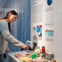 Foto eines Ausstellungsbereichs. Auf den Wänden sind Texte geschrieben, Abbildungen zu sehen und Exponate montiert. Auf einem Tisch davor stehen Lebensmittel unterschiedlicher Kategorien. Eine Frau hält einen Kassen-Scanner darüber.