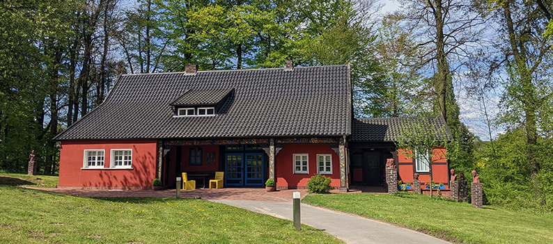 Foto des Peter-August-Böckstiegel-Haus. Das Haus ist in einem Ochsenblutrot gestrichen. Mittig ist eine blau gestrichene Flügeltür mit vielen kleinen Fenstern zu sehen. Es hat links einen kleinen Vorbau und rechts einen kleinen Anbau mit Tür.