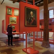 Blick in Ausstellungsraum mit Boden aus Steinplatten in verschiedenen Farben und Größen. An roten mobilen Wänden und in Vitrinen werden Gemälde, Skulpturen und andere Dinge ausgestellt. Fünf Personen schauen sich die Ausstellung an.