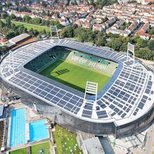 Luftaufnahme des Weserstadions. Das Stadion ist oval, die Besucherplätze überdacht. Auf dem Stadiondach stehen vier Fluchtlichtinstallationen. Links unter dem Stadion ist ein Freibad mit zwei Schwimmbecken zu erkennen. Links nks daneben fließt die Weser.
