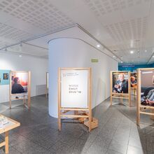Foto eines Ausstellungsbereichs im Gustav-Lübcke Museum Hamm. Auf verschiedenen Holzaufstellern sind große Fotos von älteren Menschen zu sehen. Mittig steh ein Aufsteller mit den Worten "Werde Zweit Zeug*in".
