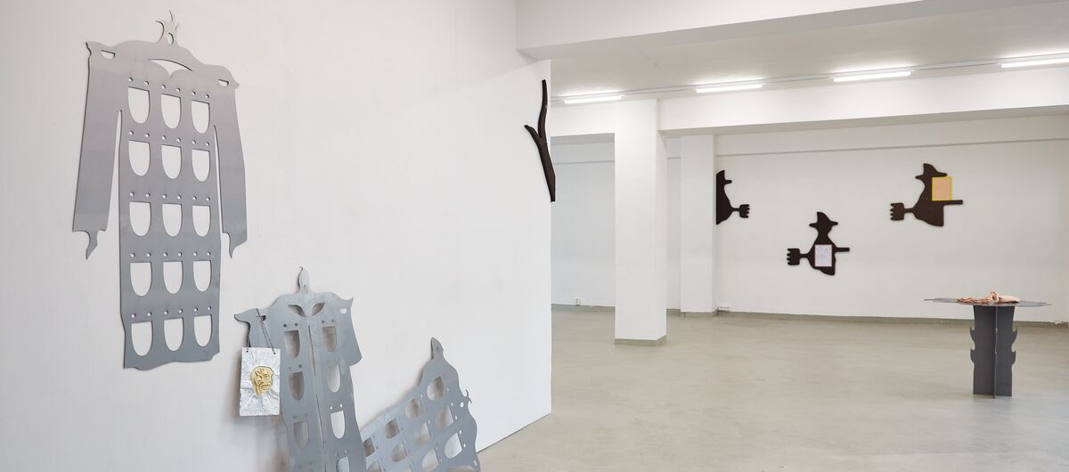 Foto eines Ausstellungsbereichs. An der linken Wand hängen und stehen drei silberne Platten in unterschiedlicher Form und systematischer Ausfräsung. An der Stirnwand sind drei schwarze Schnitte in Form einer Hexe auf einem Besen befestigt.