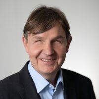Ein Mann lächelt in die Kamera. Er trägt ein hellblaues Hemd mit Kragen, darüber ein schwarzes Sakko. Seine Haare sind braun und fallen seitlich in die Stirn.