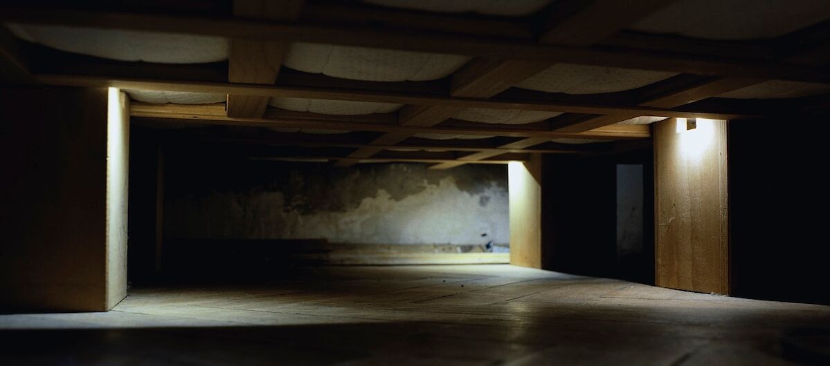 Foto in einen Raum ohne Einrichtungsgestände und Tapeten. An der Decke sind breite Holzbalken schachbrettartig angeordnet. Drei Holzbretterverschläge sind an den Seiten, an denen je eine brennende Lampe herunterstrahlt. Ansonsten ist es dunkel.