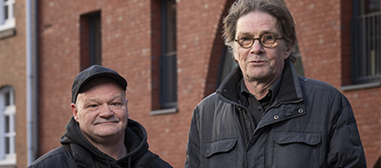 Foto von den Hinz & Kunzt Guides Chris und Uwe. Beide stehen nebeneinander vor einem Backsteinhaus, tragen warme Jacken und schauen mit einem freundlichen Blick in die Kamera..