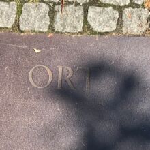 Schriftzug "Ort" eingraviert auf eine Stahlplatte, die an kleine graue Steinquadrate grenzt, welche den Bodenbelag bilden. Durch die Ritzen der Steine dringt grünes Moos.