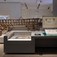Foto in einen dunkel gehaltenen Teil des Ausstellungsraums. Hell erleuchtet sind Exponate, wie z. B. ein einfacher Kutschwagen aus Holz, eine Holztruhe, eine Vitrine mit Fotos und Unterlagen, eine verziertes weißes Stofftuch.