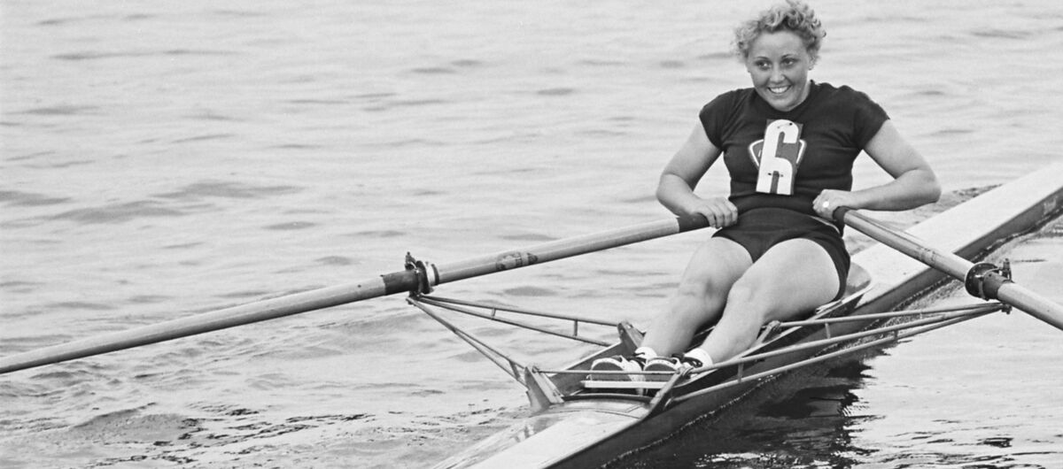 Schwarz-weiß Foto von der Ruderin Lieselotte Proll bei der Frühjahrsregatta in Grünau am 22.5.1954. Sie ist lachend auf dem Wasser am Rudern.