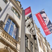 Foto der Außenansicht des Willy-Brandt-Hauses Lübeck. Die Fassade, die mit einer langen und einer kurzen Fahnen des Ausstellungshauses bestückt ist, wurde von unten schräg nach oben fotografiert. Das Haus besteht aus Sandstein.