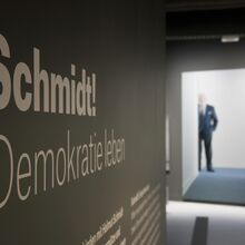 Fotos, das eine von der Seite aufgenommene Informationstafel im Vordergrund zeigt. Darauf ist deutlich "Schmidt! Demokratie leben" zu lesen. Im Hintergrund steht hinter einem Durchgang ein Mann im Anzug, der nur verschwommen zu sehen ist.