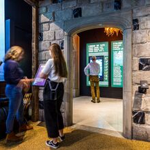 Foto in die Ausstellung. Im vorderen Raum schauen sich zwei Frauen eine Ausstellungstafel an. Durch einen Torbogen aus Stein schaut man in den nächsten Ausstellungsraum. Hier steht ein Mann vor einer Ausstellungstafel.
