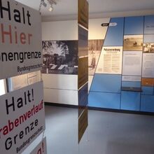 Foto eines Ausstellungsbereichs. Im Vordergrund auf der rechten Seite hängen Schilder untereinander in zwei Reihen, auf denen beispielweise steht: Halt Hier Zonengrenze Bundesgrenzschutz.