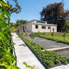Foto des Haus am Horn. Zu dem weißen bungalowartigen Gebäude mit kleinen Aufbau führen ein Kiesweg, der von wenigen Treppen unterbrochen wird., sowie ein barrierefreier betonierter Weg. Drumherum sind grüne Hecken, Beete, Bäume und eine Rasenfläche.