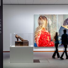 Foto der Ausstellungsansichten zu „Zerreißprobe. Kunst zwischen Politik und Gesellschaft. Sammlung der Nationalgalerie 1945-2000“. In einer Vitrine steht ein großer Damenschuh auf einem Sockel. Dahinter hängt ein großes Foto von zwei Frauen im Bad.