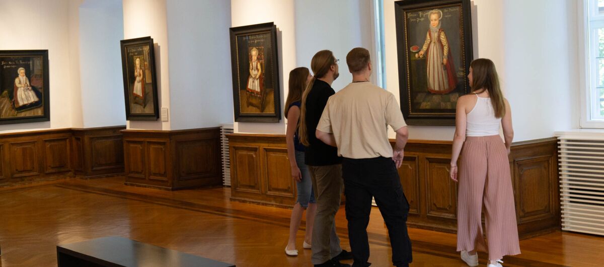 Blick in Ausstellungsraum mit Boden und hüfthohen Wandvertäfelungen aus Holz. Vier Gemälde hängen an den Wänden. Um das ganz rechts im Bild stehen zwei Frauen und zwei Männer und betrachten es. Links unten steht eine schwarze minimalistische Besucherbank.