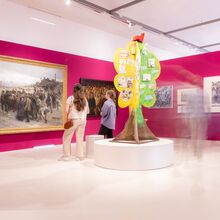 Foto eines Ausstellungsbereichs. An einer roten Wand hängen große und kleine Gemälde, die vornehmlich Menschengruppen abbilden. Zwei Frauen sehen sich die Bilder an. Hinter ihnen steht auf einem runden Podest ein gebastelter Baum.