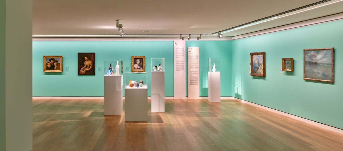 Foto eines Ausstellungsbereichs. An einer helltürkiesen Wand hängen in einer Reihe verschiedenste alte Gemälde in Rahmen. Davor stehen vier schmale Vitrinen, in denen sich Gefäße befinden.