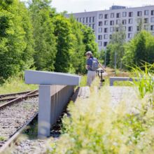 Blick aus Kniehöhe. Links sind zwei Gleise zu sehen. Mittig eine lange Reihe von Informationstafeln aus Beton, die von einem Menschen mit Fahrrade betrachtet werden. Im Hintergrund sind grüne Bäume sowie eine Reihenhauszeile zu sehen.